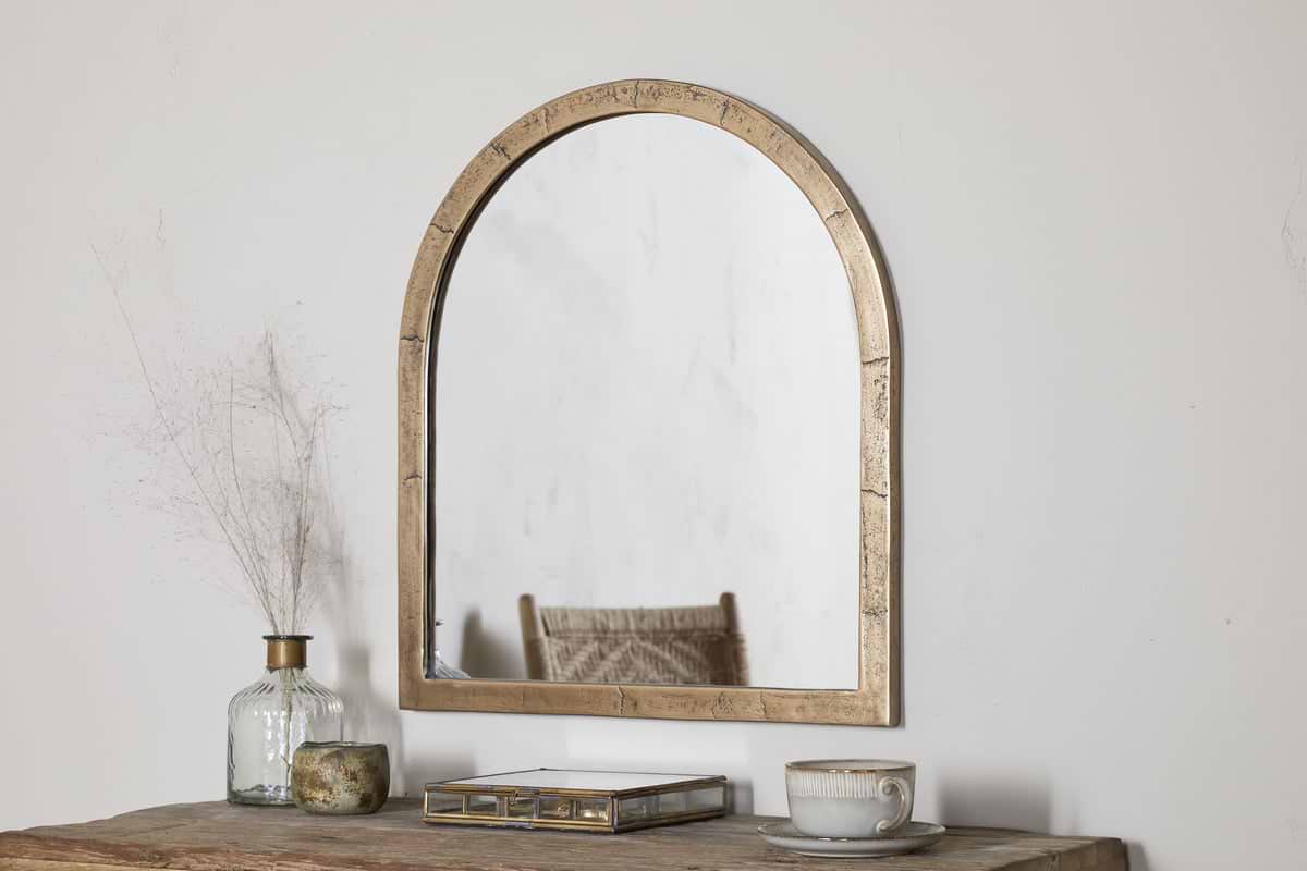 A Murwara Wall Mirror by nkuku in Antique Brass hangs on a white wall above a rustic wooden table topped with a glass vase of dried branches, a decorative box, a candle, and a cup with saucer.