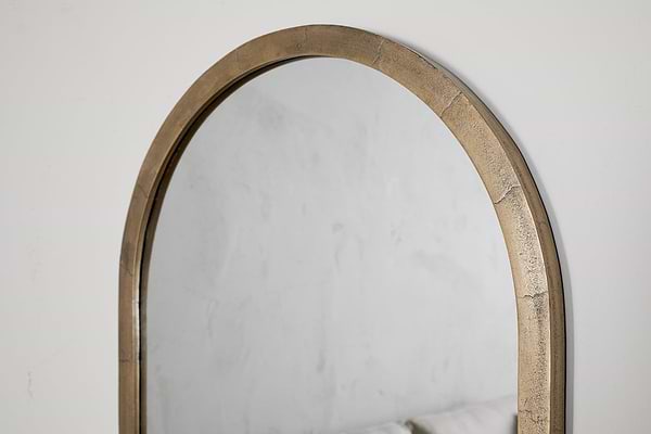 A close-up of the nkuku Murwara Wall Mirror in Antique Brass, reflecting a light-textured wall and the upper edge of a pale sofa.