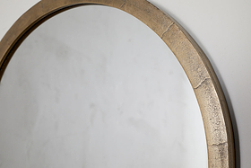 The Murwara Wall Mirror by nkuku features a round, antique brass frame with a textured, metallic gold finish, shown hanging on a plain, light-colored wall and reflecting part of the surface.