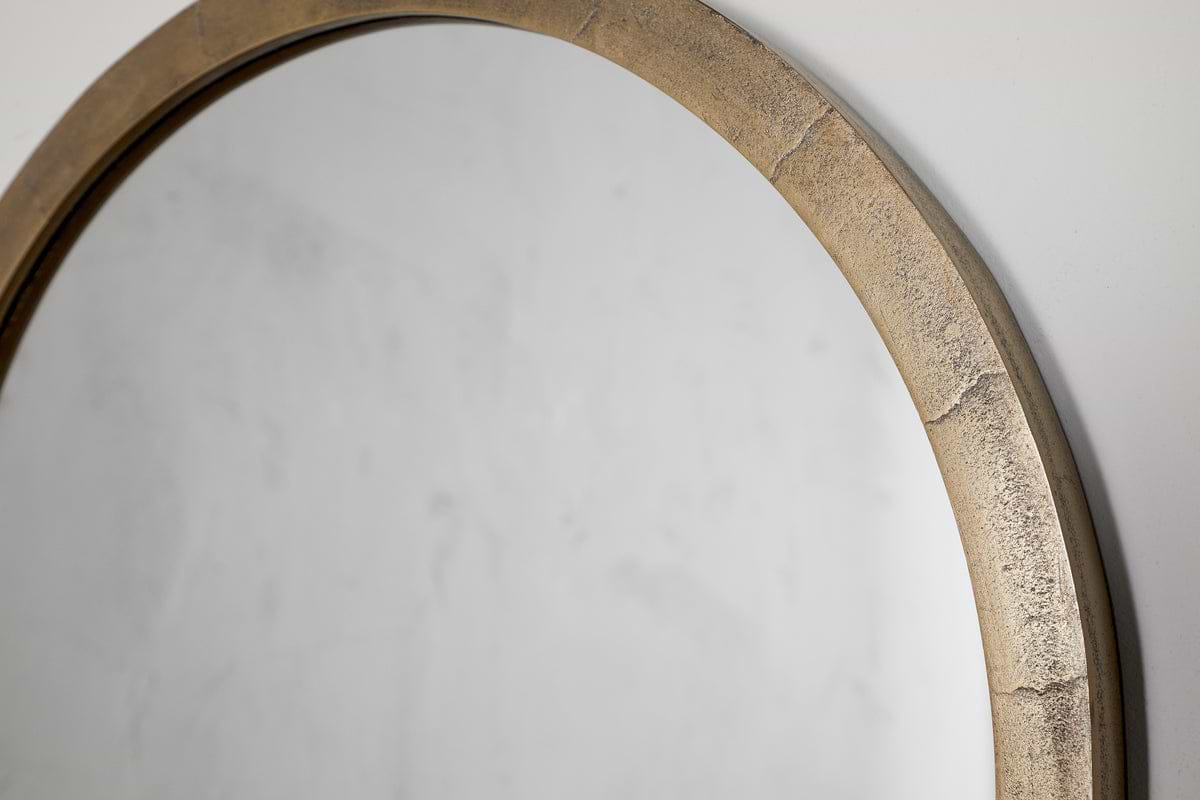 The Murwara Wall Mirror by nkuku features a round, antique brass frame with a textured, metallic gold finish, shown hanging on a plain, light-colored wall and reflecting part of the surface.