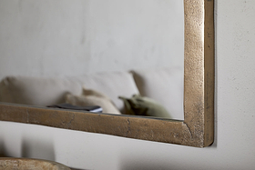 A close-up of the nkuku Murwara Wall Mirror in Antique Brass reflects part of a beige sofa with a light blanket and green cushion, set against a neutral wall.