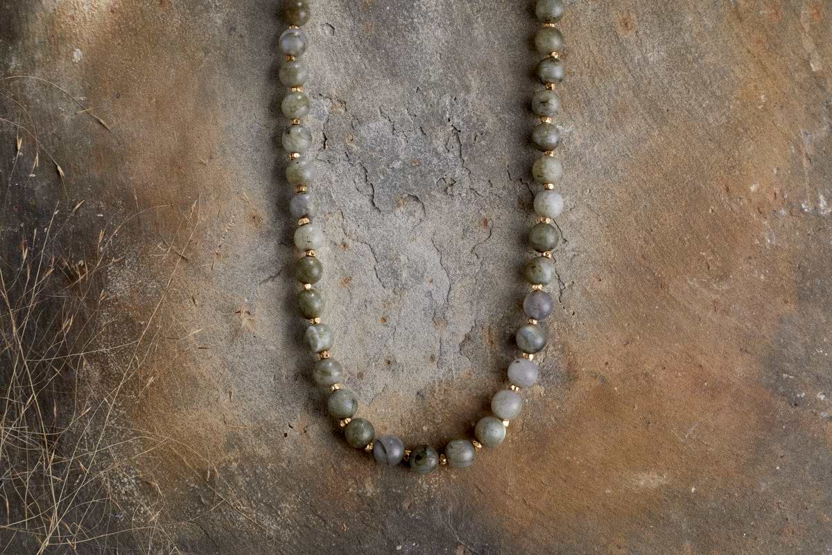 The Marai Necklace - Labradortite by nkuku features round, earthy labradorite beads arranged in a semicircle, displayed on a rustic brown textured surface for an elegant look.