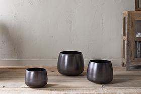 Three nkuku Maliki Plant Pots in aged bronze with a matte finish, each a different height, are arranged on a wooden floor by a light beige wall. A rustic shelf with books is visible to the right, complementing the minimalist look.
