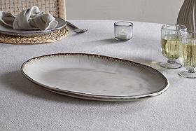 The nkuku Malia Large Oval Platter in cream, featuring an organic shape and textured rim, rests on a light gray tablecloth beside two pale drinks, a lit candle, and a place setting with plate, napkin, and woven placemat.