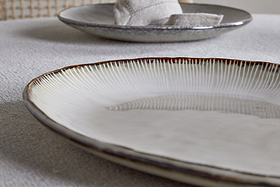 A close-up of the nkuku Malia Large Oval Platter in cream, featuring a textured, ridged rim and organic shape on a light tablecloth, with another platter and a folded napkin blurred in the background.
