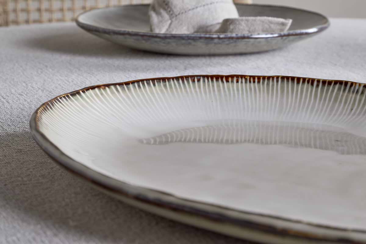A close-up of the nkuku Malia Large Oval Platter in cream, featuring a textured, ridged rim and organic shape on a light tablecloth, with another platter and a folded napkin blurred in the background.