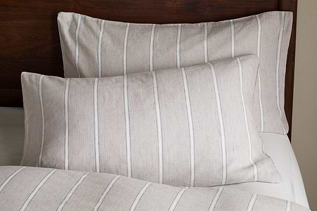 Two Mora Hemp & Cotton Housewife Pillowcases in Stone Grey by nkuku, featuring vertical white stripes, rest on a bed with matching bedding against a dark wooden headboard.