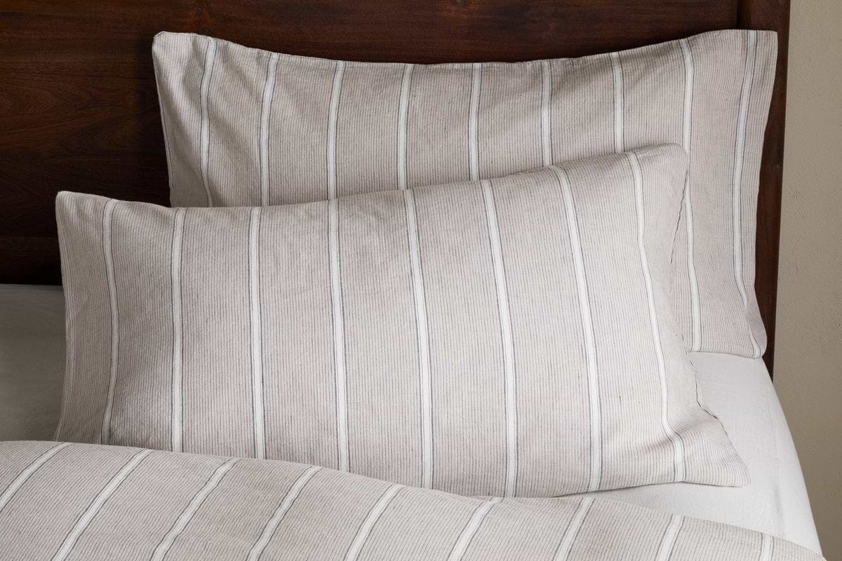 Two Mora Hemp & Cotton Housewife Pillowcases in Stone Grey by nkuku, featuring vertical white stripes, rest on a bed with matching bedding against a dark wooden headboard.