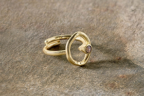The Mari Labradorite Ring - Gold from nkuku features a hypoallergenic, gold-plated adjustable open band with an irregular circular design and small labradorite gemstone, displayed on a textured stone surface.