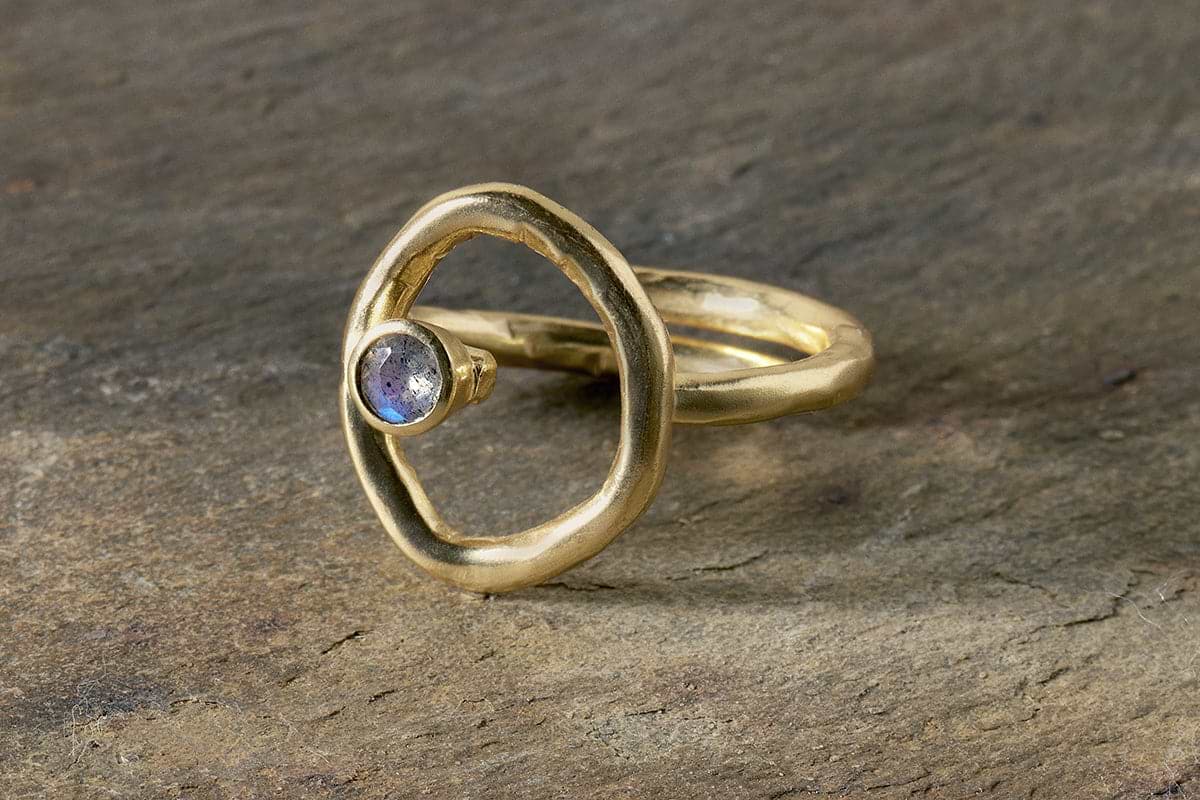 The nkuku Mari Labradorite Ring - Gold features a gold plated, organic open circle design with a bezel-set iridescent Labradorite stone, a matte hypoallergenic finish, and is shown on textured stone.