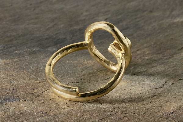 The Mari Labradorite Ring - Gold by nkuku features a hand-shaped, irregular band with a rough, organic texture, giving it an artisan feel. It rests on an earthy-toned, textured stone surface that highlights its sculptural quality.