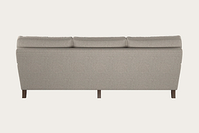 A beige Marri Super Grand Sofa - Brera Linen Natural by Nkuku is shown from the back, highlighting its modern, minimalist design with clean lines, a cushioned backrest, and short dark wooden legs against an off-white background. Handcrafted in a Nottinghamshire workshop with premium Brera linen upholstery, this three-seater sofa offers contemporary elegance.
