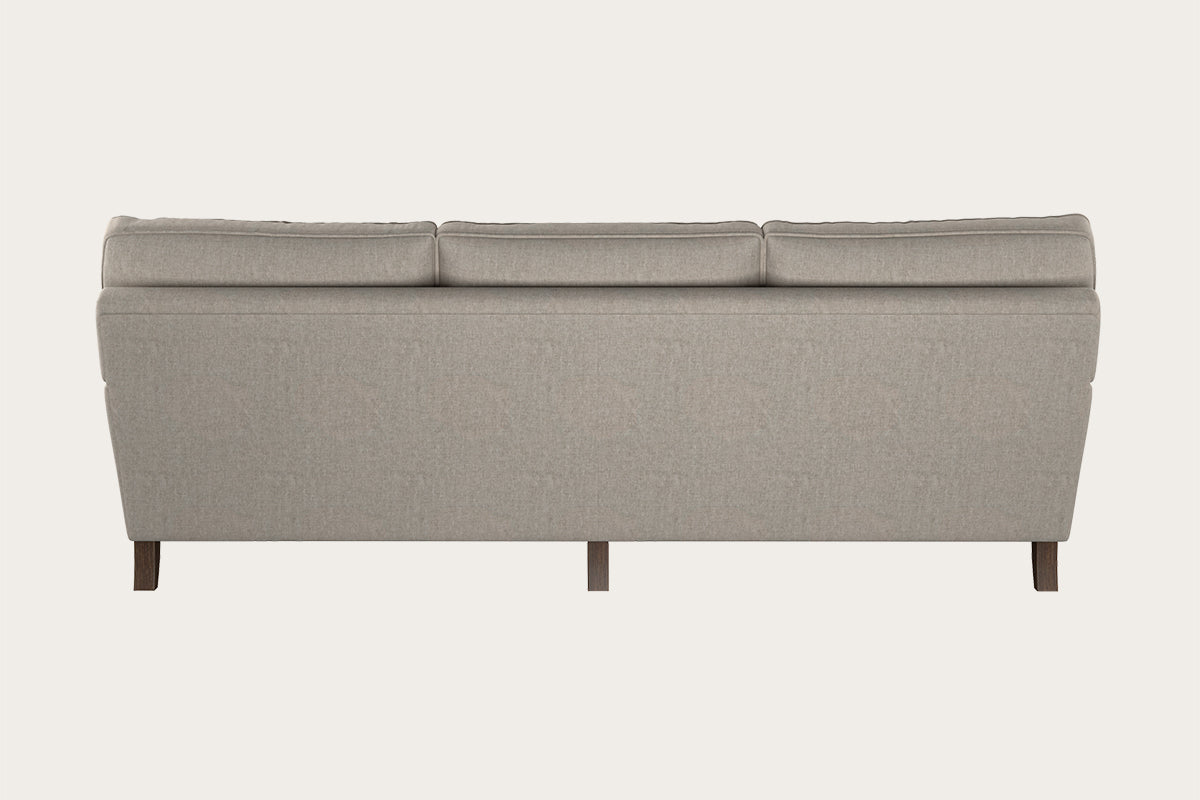 A beige Marri Super Grand Sofa - Brera Linen Natural by Nkuku is shown from the back, highlighting its modern, minimalist design with clean lines, a cushioned backrest, and short dark wooden legs against an off-white background. Handcrafted in a Nottinghamshire workshop with premium Brera linen upholstery, this three-seater sofa offers contemporary elegance.