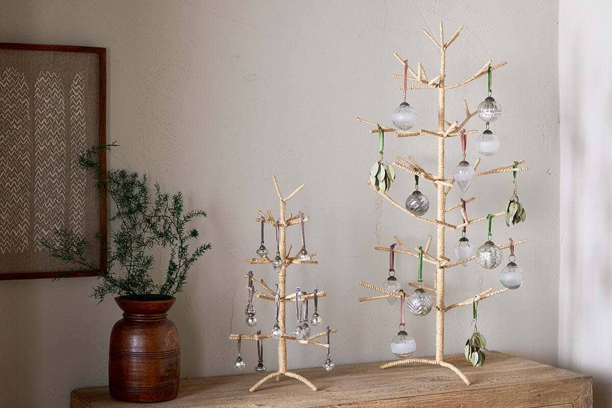 Two nkuku Mala Jute Trees in natural, with gold and woven jute branches, stand on a wooden surface. Each is adorned with glass ornaments and green accents, complemented by a brown vase of pine foliage and framed textile art for a festive look.