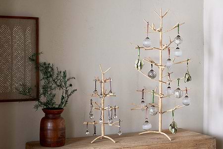 Two nkuku Mala Jute Trees in natural, with gold and woven jute branches, stand on a wooden surface. Each is adorned with glass ornaments and green accents, complemented by a brown vase of pine foliage and framed textile art for a festive look.