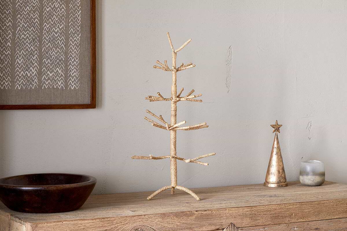 A minimalist gold wire tree sculpture stands on a rustic wooden surface beside nkuku’s Mala Jute Tree - Natural, a festive metallic cone with star topper, candle, and dark wooden bowl. The backdrop is a neutral wall with framed woven jute branches art.