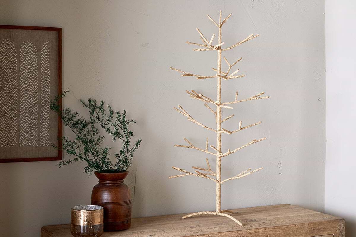 The Mala Jute Tree - Natural by nkuku stands on a wooden table beside a brown ceramic vase with green foliage, a decorative jar, and framed textile art, creating a festive display against the pale wall.