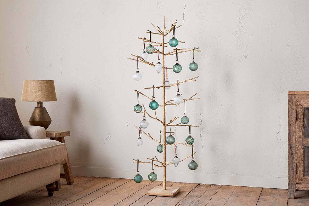 The nkuku Mala Base Jute Tree - Natural, with minimalist jute branches and gold accents, is adorned with green and clear glass ornaments, standing beside a beige sofa, rustic wooden cabinet, and table lamp against an off-white wall.