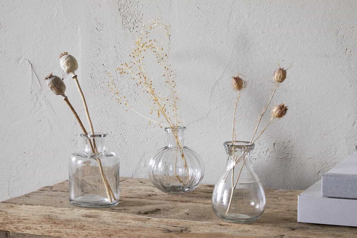 The Mayumi Recycled Glass Bud Vases - Clear (Set of 3) by nkuku are displayed with dried stems on a rustic wooden surface beside two stacked gray books, set against a textured light gray wall.