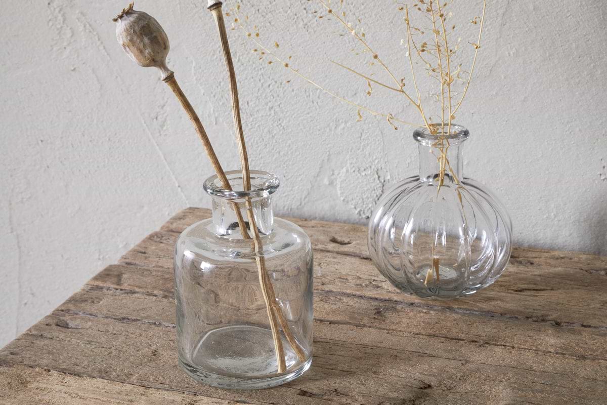 Three Mayumi Recycled Glass Bud Vases by nkuku sit on a rustic wooden surface, each holding dried botanicals. The clear glass vases are set against a textured, light-colored wall.