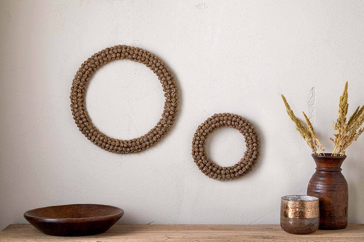 Two Madhi Wire Ball Wreaths by nkuku in brass create a striking sculptural decor display on a light beige wall. Below, a wooden bowl, brown vase with dried grasses, and metallic container complete the natural, rustic arrangement.