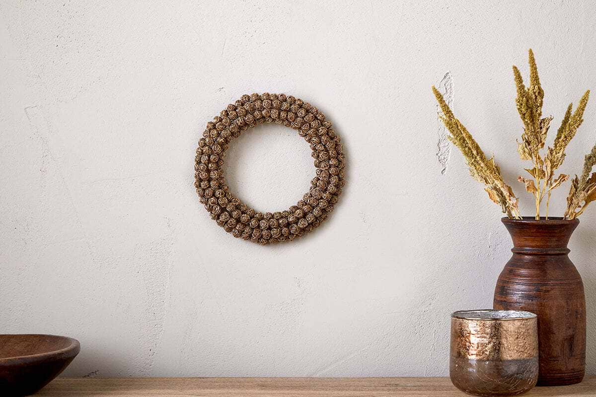 A Madhi Wire Ball Wreath in brass by nkuku hangs on a textured off-white wall. Below, a dark wooden bowl, brown vase with dried grasses, and a metallic candle holder create a calm, natural display.