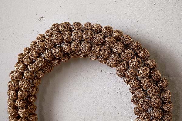 A close-up of the Madhi Wire Ball Wreath - Brass by nkuku, featuring tightly arranged brass wire balls in a layered design, elegantly displayed on a textured off-white wall.