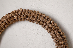 A close-up shows the Madhi Wire Ball Wreath in brass by nkuku, featuring tightly woven wire balls. Part of the arc is visible against a textured light wall, creating a striking sculptural display.