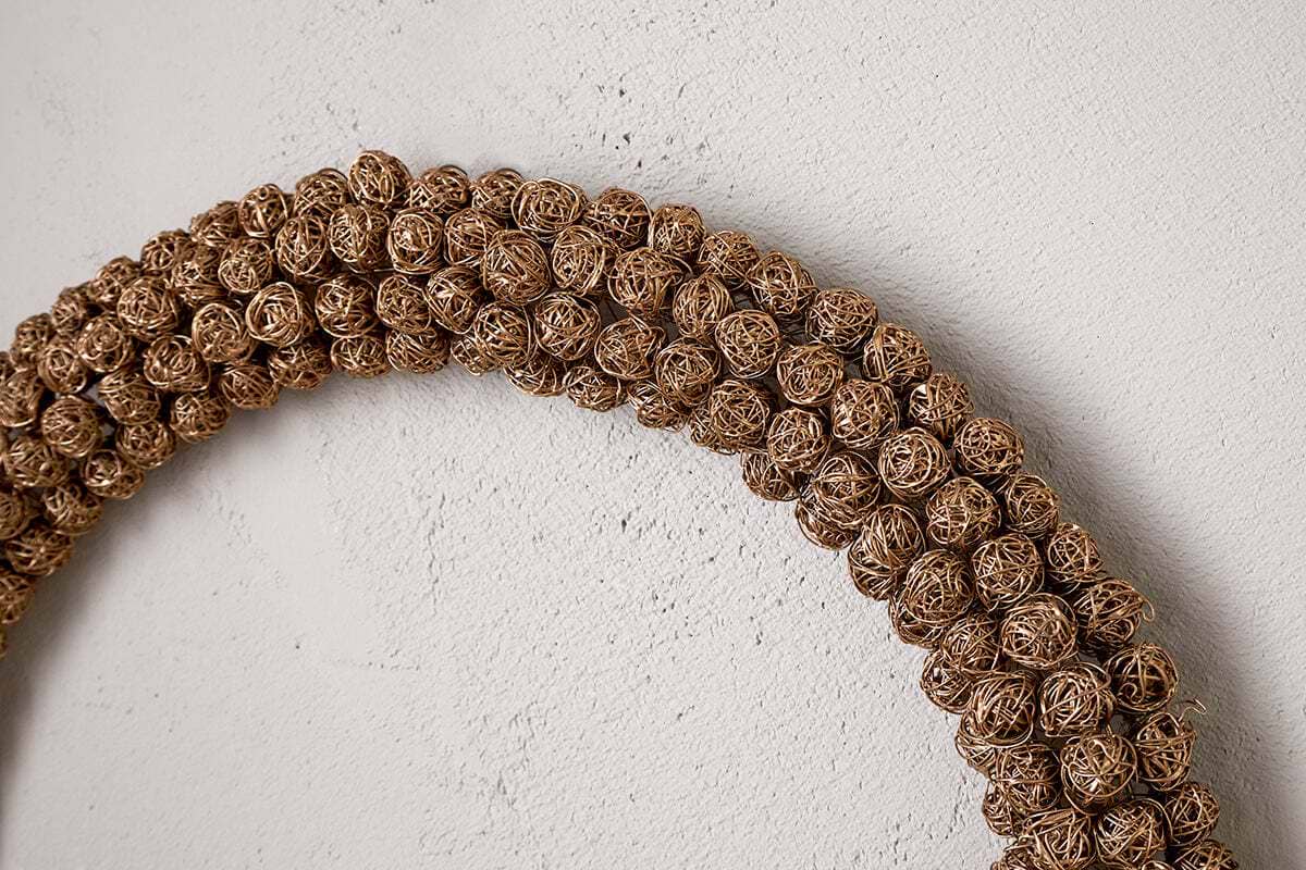 A close-up shows the Madhi Wire Ball Wreath in brass by nkuku, featuring tightly woven wire balls. Part of the arc is visible against a textured light wall, creating a striking sculptural display.