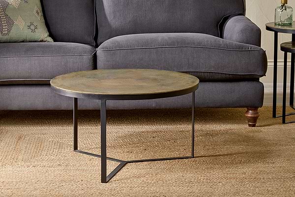 The nkuku Maba Coffee Table, featuring a bronze top and slim black legs, stands on a beige woven rug before a dark gray sofa with wooden legs and a green cushion. A small round side table is partly visible on the right.