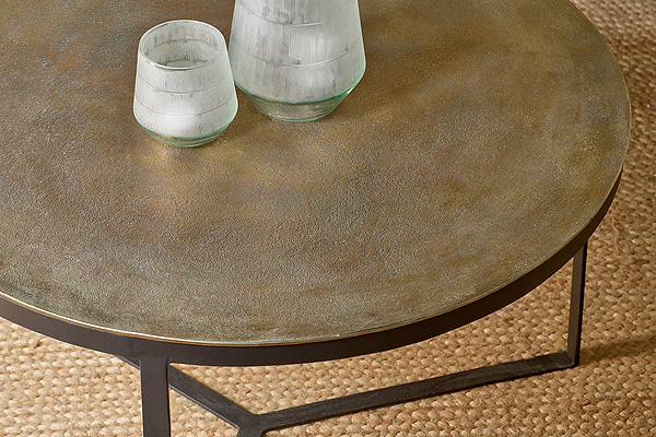 The Maba Coffee Table by nkuku features a textured bronze top and slender black metal frame, shown here with two frosted glass cups and a carafe on a woven beige rug, adding style and warmth to your living room.