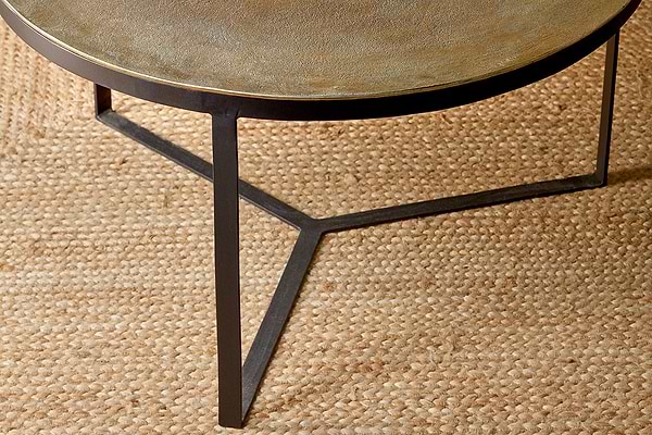 Close-up of the Maba Coffee Table by nkuku featuring a matte, subtly golden antique brass round top and a geometric dark three-legged metal base, shown on a beige jute rug.