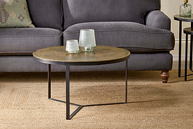 The Maba Coffee Table by nkuku, featuring a metallic top and black geometric legs, sits on a woven rug before a gray sofa. Two glass vases and a patterned pillow complete this charming and inviting living room setup.