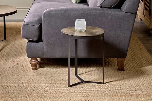 The Maba Nesting Side Table by nkuku, featuring a thin metal base, stands beside a dark gray sofa with wooden legs and curved armrests on a woven beige rug. A glass of water rests on the table, with part of a wooden cabinet in the background.