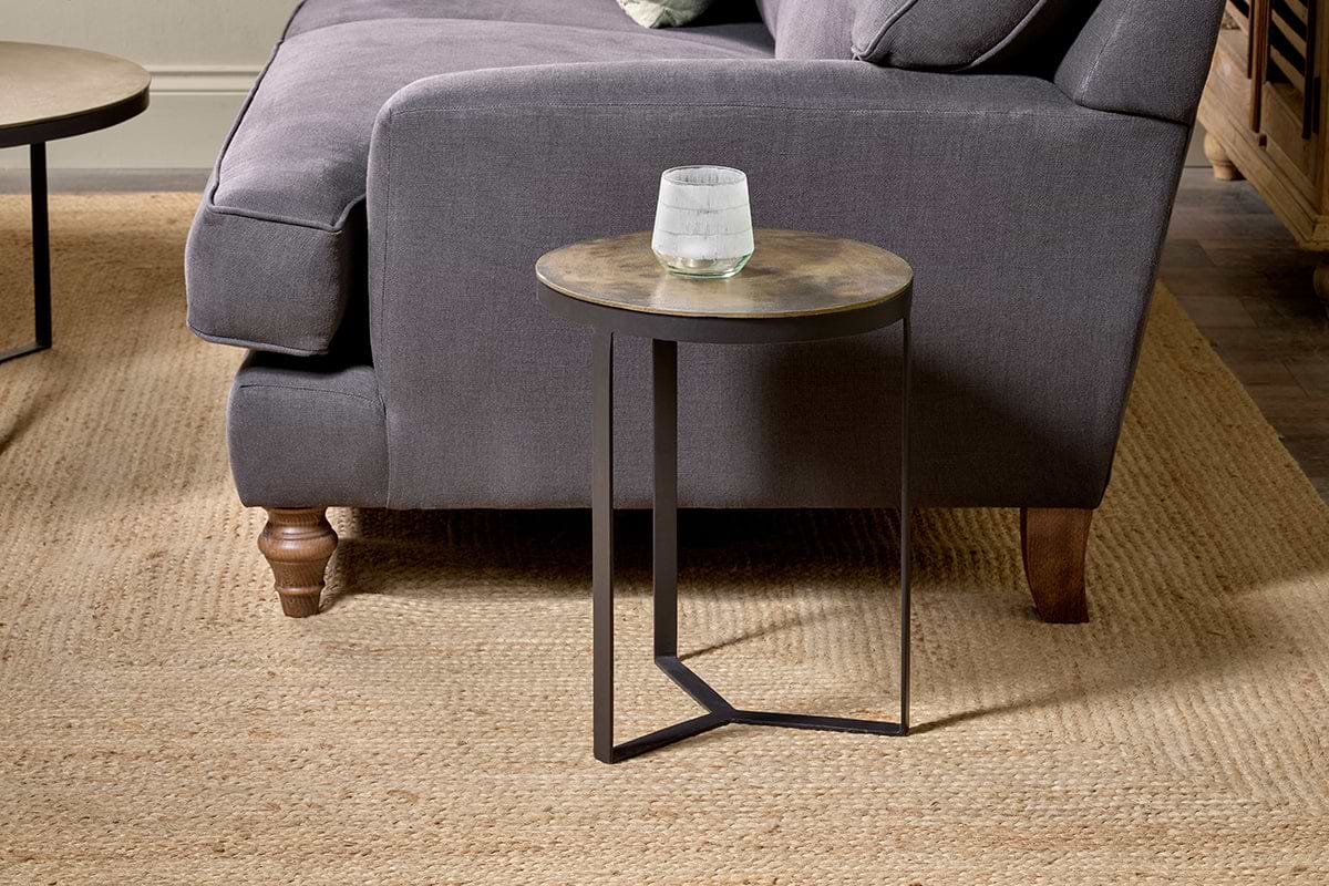 The Maba Nesting Side Table by nkuku, featuring a thin metal base, stands beside a dark gray sofa with wooden legs and curved armrests on a woven beige rug. A glass of water rests on the table, with part of a wooden cabinet in the background.
