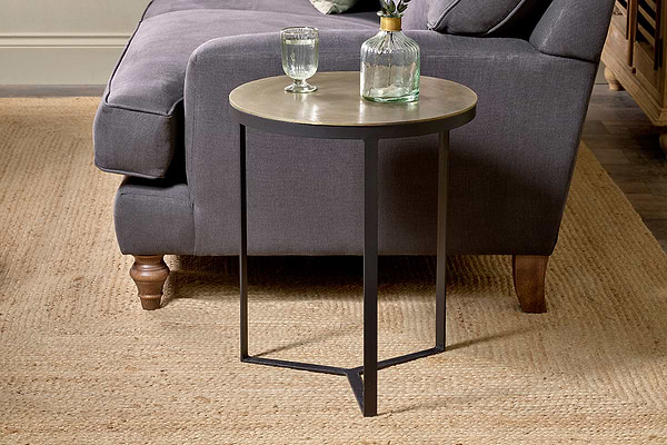 A round Maba Nesting Side Table by nkuku with a metal frame and Maba tabletop stands on a woven beige rug beside a dark gray upholstered sofa. A clear glass and decanter filled with water rest atop it, with wooden flooring and furniture in the background.