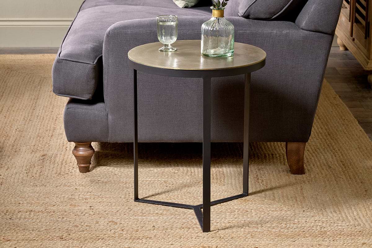 A round Maba Nesting Side Table by nkuku with a metal frame and Maba tabletop stands on a woven beige rug beside a dark gray upholstered sofa. A clear glass and decanter filled with water rest atop it, with wooden flooring and furniture in the background.