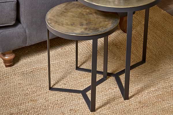 The nkuku Maba Nesting Side Tables, featuring round tabletops and black metal legs, overlap on a beige woven rug beside a gray sofa with wooden legs, showcasing their compact, space-saving design.