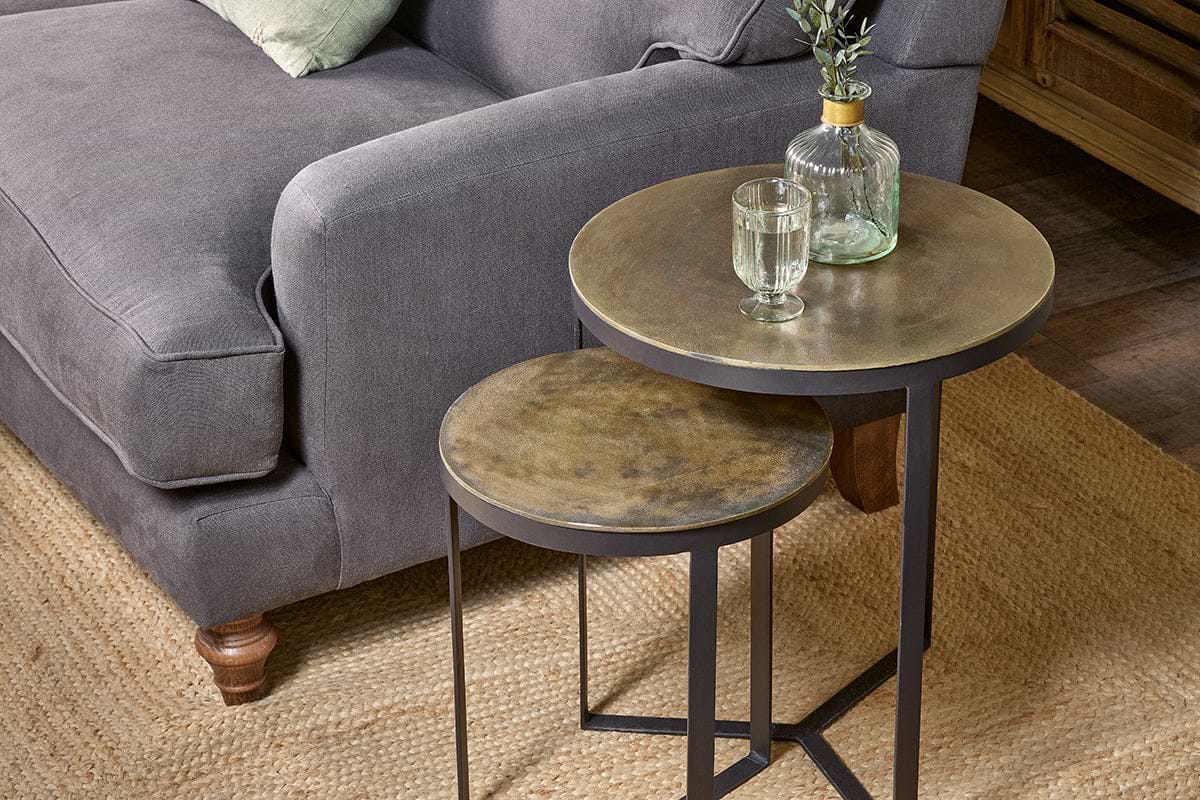 A gray sofa with a cushion is next to two nkuku Maba Nesting Side Tables with brass tops. On the larger table are a glass of water and a clear bottle with green foliage. All rest on a woven jute rug over wooden floors.