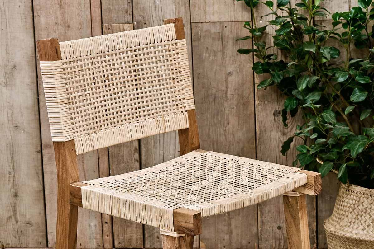The Madrisana Acacia & Rattan Bar Stool by nkuku, featuring a woven light-colored seat and backrest, is placed before rustic wooden paneling. A potted green plant appears partially on the right side of the image.