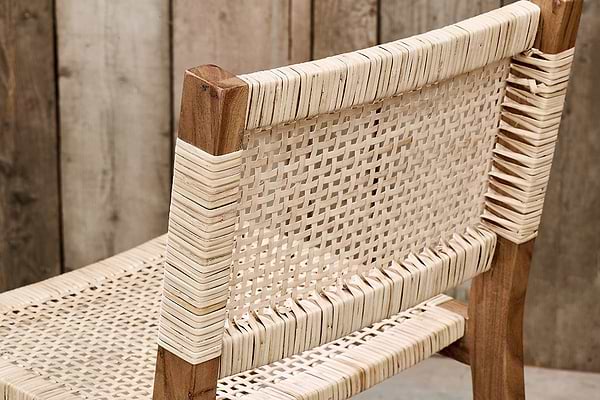 A close-up of the Madrisana Acacia & Rattan Bar Stool by nkuku, featuring a woven cane seat and backrest on a light brown acacia frame. Wrapped cane details accent the joints, set against rustic, vertically arranged wooden planks in the background.