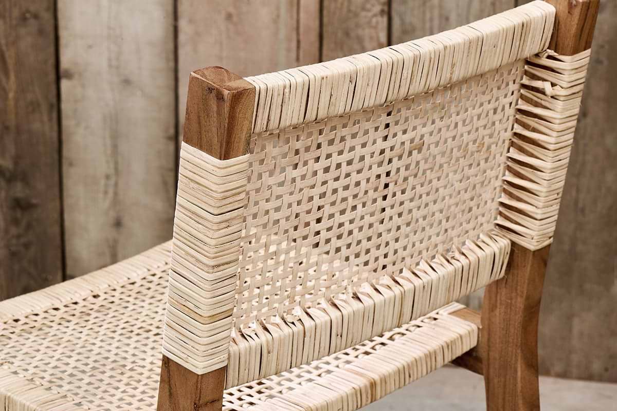 A close-up of the Madrisana Acacia & Rattan Bar Stool by nkuku, featuring a woven cane seat and backrest on a light brown acacia frame. Wrapped cane details accent the joints, set against rustic, vertically arranged wooden planks in the background.
