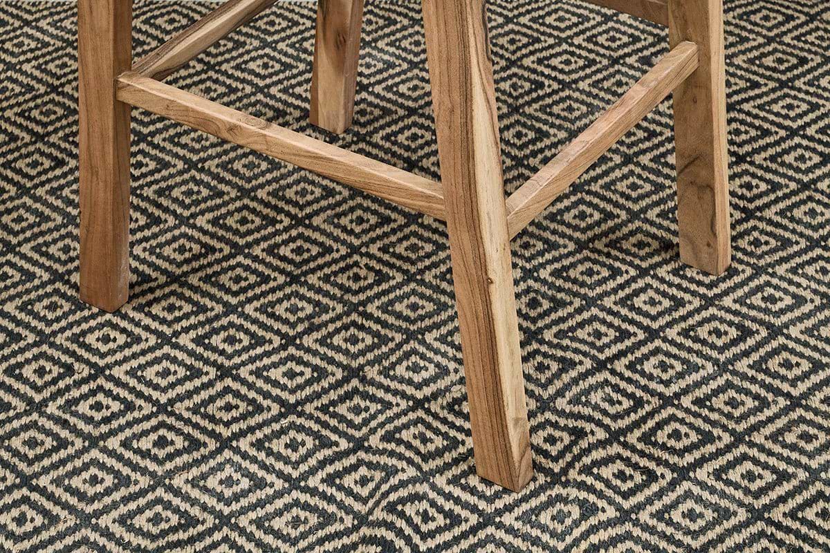 Close-up of the Madrisana Acacia & Rattan Bar Stool by nkuku, showing its natural-finish legs on a black and beige diamond-patterned woven rug—highlighting the contrast between the stool’s sturdy frame and the geometric design below.