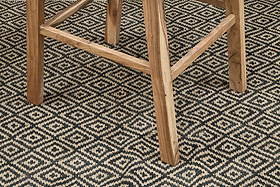 A close-up of the Madrisana Acacia & Rattan Bar Stool by nkuku shows its light acacia legs on a woven black and beige rug, the diamond pattern contrasting with the smooth natural wood grain.