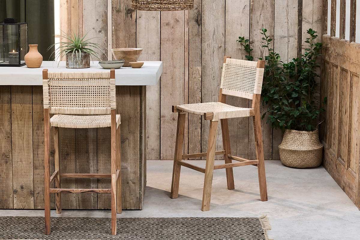 Two Madrisana Acacia & Rattan Bar Stools by nkuku, with woven seats and backs, sit beside a rustic wood-paneled counter. A potted green plant in a woven basket adds to the cozy, minimalist space with natural tones.