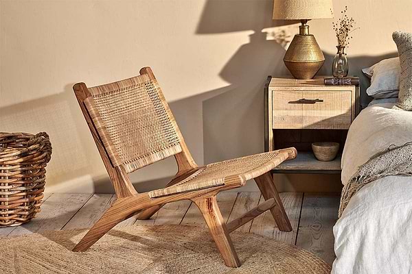 The Madrisana Acacia & Rattan Woven Chair by nkuku sits on a jute rug, next to a bedside table topped with a lamp and accompanied by a basket.