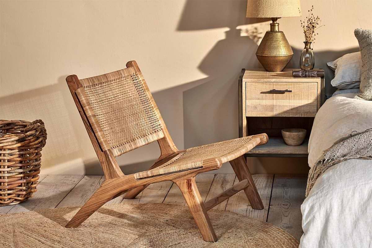 The Madrisana Acacia & Rattan Woven Chair by nkuku sits on a jute rug, next to a bedside table topped with a lamp and accompanied by a basket.