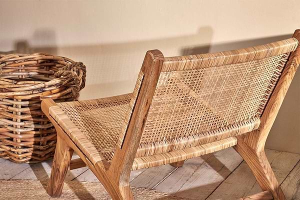 The Madrisana Acacia & Rattan Woven Chair by nkuku sits low on a light wooden floor next to a large round wicker basket, casting a soft shadow on a beige wall for a warm, inviting atmosphere.