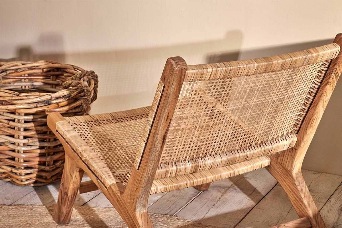 The Madrisana Acacia & Rattan Woven Chair by nkuku sits low on a light wooden floor next to a large round wicker basket, casting a soft shadow on a beige wall for a warm, inviting atmosphere.
