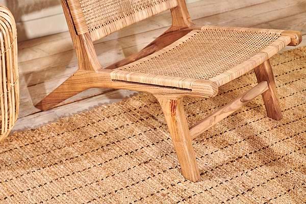 A close-up of the nkuku Madrisana Acacia & Rattan Woven Chair sits on a textured natural fiber rug. Warm, neutral tones and sunlight cast soft shadows on the light wood floor, enhancing the cozy minimalist vibe.